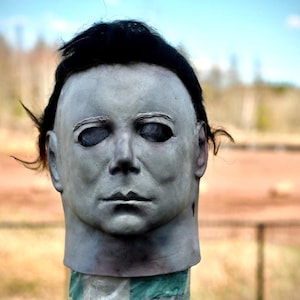 May include: A pale grey, full-head mask featuring a stoic facial expression, deep-set dark eye sockets, and moulded dark brown hair. The mask has a smooth, matte finish with subtle shading around the nose, mouth, and jawline, creating a realistic appearance.