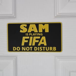 May include: A rectangular black and yellow sign with the text "SAM IS PLAYING FIFA DO NOT DISTURB" is mounted on a white door. The sign has a yellow border and the text is in a bold, yellow font.