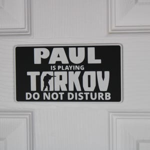 May include: A rectangular black and white sign with the text "PAUL IS PLAYING TARKOV DO NOT DISTURB" in bold white letters. The sign is mounted on a white door with paneling.