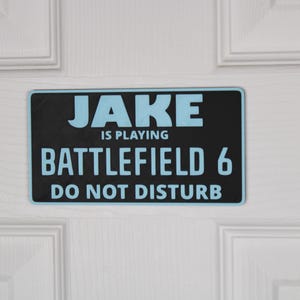 Pode incluir: Uma placa retangular preta com o texto "JAKE IS PLAYING BATTLEFIELD 6 DO NOT DISTURB" em letras azul claro. A placa está montada em uma porta branca com painéis.