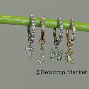 Könnte beinhalten: Vier Creolen-Ohrringe mit Anhängern. Die Anhänger sind in Form der Buchstaben T, J, W und K sowie eines Kreuzes gehalten. Die Creolen und Anhänger sind silber- und goldfarben und mit kleinen, klaren Steinen besetzt. Der Text "@Dewdrop Macket" befindet sich unten.