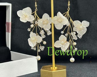 May include: Elegant floral earrings with white flower accents, pearl beads, and crystal embellishments. The earrings are suspended from gold-colored hooks and are displayed on a gold stand. The earrings have a delicate, feminine design.