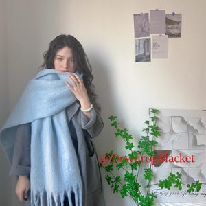 May include: A light blue scarf with fringe is draped over a person wearing a gray coat. The scarf is made of a soft, textured material. The image also shows a plant with green leaves and the text "@DewdropMarket" and "Enjoy your life every day".