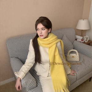 May include: A woman in a cream suit and a vibrant yellow scarf with a fringed edge. She is seated on a striped sofa, with a small white handbag. The scarf is wrapped around her neck and shoulders.