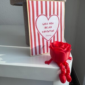 Könnte beinhalten: Eine Valentinstagskarte mit rot-weißen Streifen und einem Herz mit der Aufschrift "Willst du mein Valentin Emma sein". Eine rote Rosenfigur mit Armen und Beinen liegt neben der Karte.