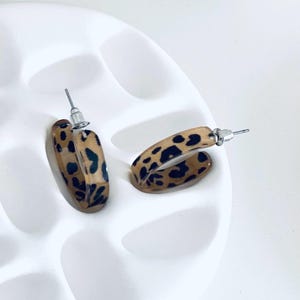 Könnte beinhalten: Zwei ovale Creolen-Ohrringe mit einem braunen und schwarzen Leopardenmuster. Die Ohrringe liegen auf einer weißen Oberfläche. Die Ohrringe haben silberfarbene Stifte.