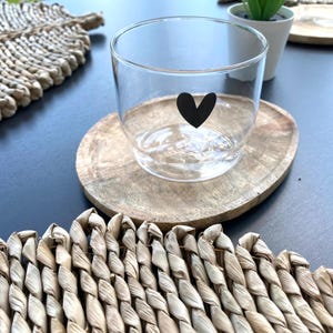 Op de afbeelding: Een helder glazen tumbler met een zwart hartontwerp, rustend op een ronde houten onderzetter. De onderzetter staat op een donkere ondergrond, met geweven placemats op de voorgrond en een kleine plant in een pot op de achtergrond. Het glas is leeg.