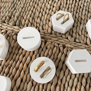 Könnte beinhalten: Eine Auswahl an weißen, geometrischen Ringhaltern mit goldenen Ringen. Die Ringe, darunter ein geflochtener Bandring und ein Wellenring, werden auf runden und sechseckigen Haltern präsentiert. Der Hintergrund ist ein geflochtenes Naturfasergewebe.