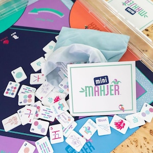 May include: A close-up of a game board with colorful tiles and a blue cloth bag. The game board is labeled "mini MAHJER" and features a jester design. The tiles are numbered and have various symbols and designs.