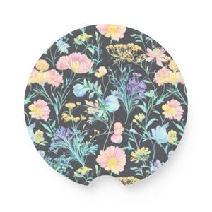 May include: A round car coaster with a floral pattern. The design features pastel-colored flowers in pink, yellow, and blue against a dark gray background. The coaster has a small notch on one side.