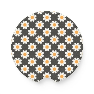 May include: A round car coaster with a daisy pattern. The coaster has a dark gray background with a repeating pattern of white daisies with orange centers. The coaster has a small notch on one side.