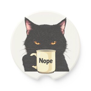 May include: A round white ceramic coaster featuring a graphic illustration of a black cat with orange eyes holding a cream-colored mug. The mug has the word Nope printed in black letters on the front. The coaster has a small notch at the bottom.