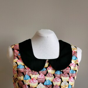 K&ouml;nnte beinhalten: Ein &auml;rmelloses Kleid mit einem schwarzen Peter-Pan-Kragen. Das Kleid ist mit einem Muster aus bunten Cupcakes mit verschiedenen Frostings und Streuseln bedeckt.