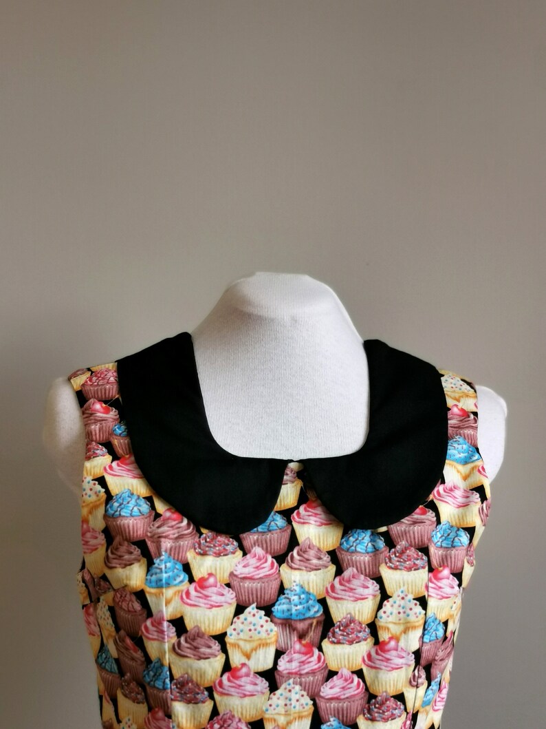 K&ouml;nnte beinhalten: Ein &auml;rmelloses Kleid mit einem schwarzen Peter-Pan-Kragen. Das Kleid ist mit einem Muster aus bunten Cupcakes mit verschiedenen Frostings und Streuseln bedeckt.