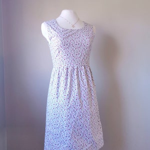 Peut inclure: Une robe blanche sans manches avec un petit motif floral dans les tons de violet et de vert. La robe a un col rond et un corsage ajust&eacute;. La jupe est l&eacute;g&egrave;rement fronc&eacute;e &agrave; la taille et arrive au niveau du genou.