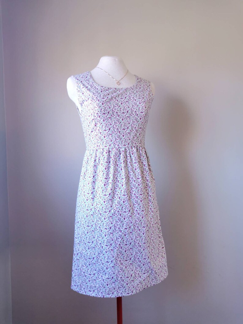 Peut inclure: Une robe blanche sans manches avec un petit motif floral dans les tons de violet et de vert. La robe a un col rond et un corsage ajust&eacute;. La jupe est l&eacute;g&egrave;rement fronc&eacute;e &agrave; la taille et arrive au niveau du genou.