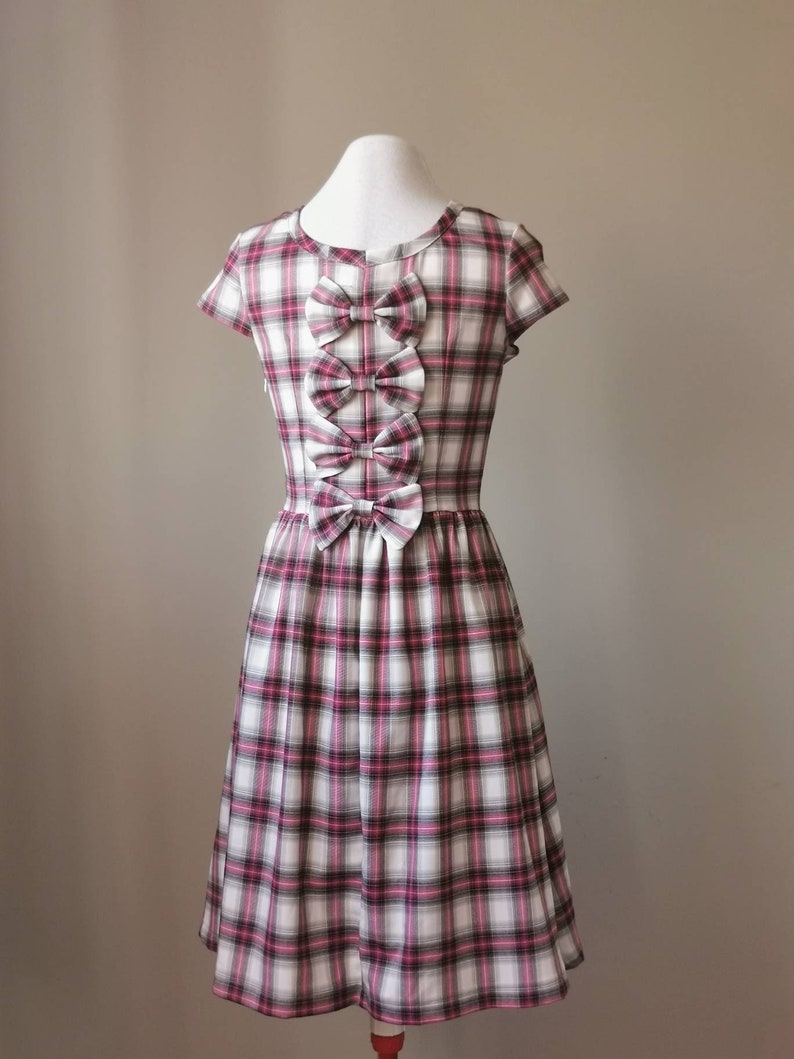 Puede incluir: Un vestido de cuadros blancos y rosas con mangas cortas y una fila de lazos en la espalda.