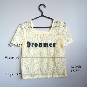 May include: A pale yellow lace top with a scoop neck and short sleeves. The top has the word "Dreamer" printed on the front. The top measures 35 inches in bust, 35 inches in waist, 36 inches in hips, and 18.5 inches in length.