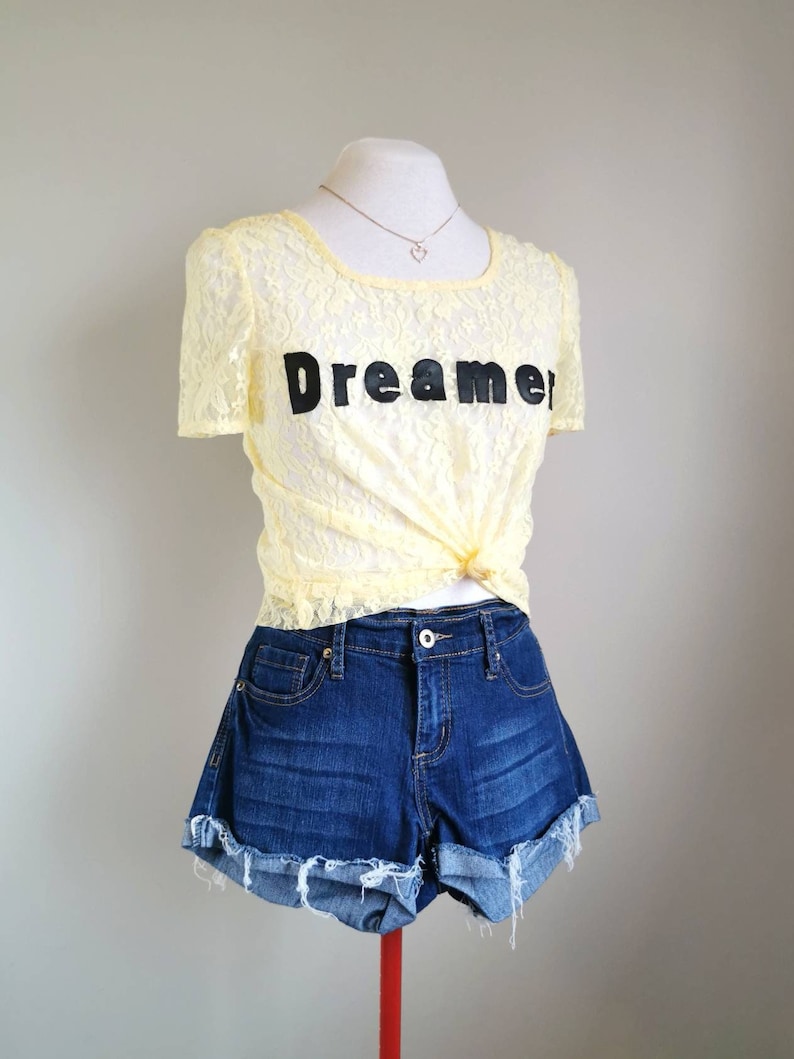 May include: A yellow lace top with short sleeves and the word "Dreamer" printed on the front. The top is paired with blue denim shorts.
