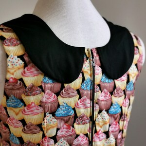 K&ouml;nnte beinhalten: Ein &auml;rmelloses Kleid mit einem schwarzen Peter-Pan-Kragen und einem Rei&szlig;verschluss. Das Kleid ist mit einem Muster aus bunten Cupcakes bedeckt.