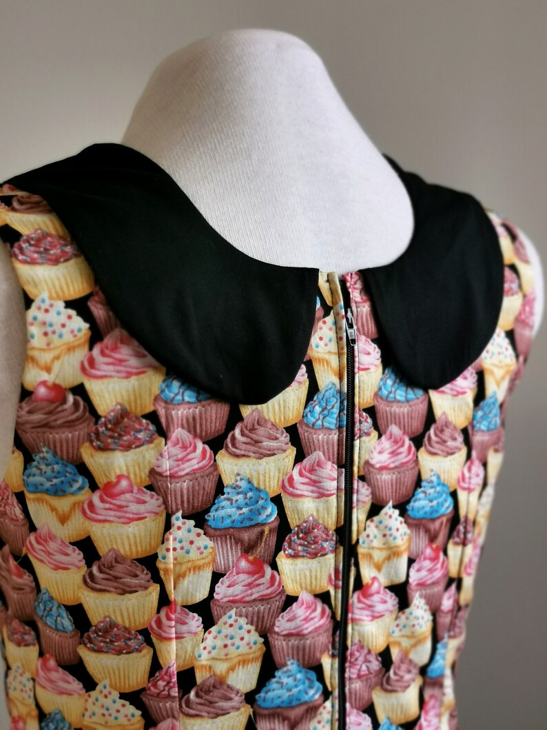 K&ouml;nnte beinhalten: Ein &auml;rmelloses Kleid mit einem schwarzen Peter-Pan-Kragen und einem Rei&szlig;verschluss. Das Kleid ist mit einem Muster aus bunten Cupcakes bedeckt.