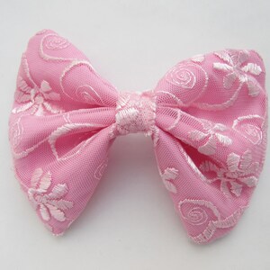 May include: A pink bow with white floral embroidery. The bow is made of a soft, sheer fabric.