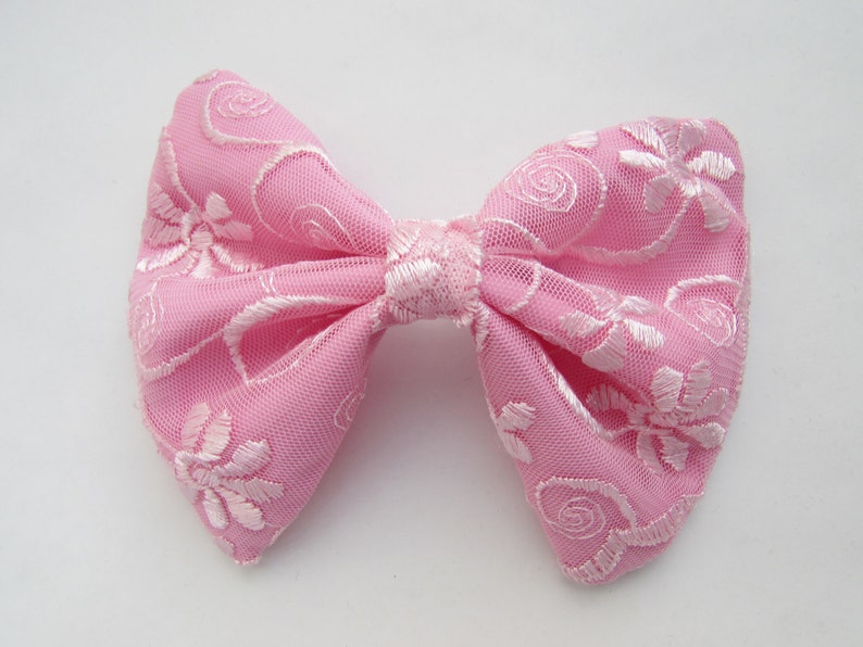 May include: A pink bow with white floral embroidery. The bow is made of a soft, sheer fabric.