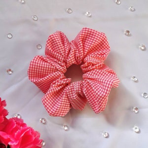 May include: A pink and white gingham hair scrunchie. The scrunchie is made of fabric and is tied in a knot.