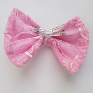 May include: A pink lace bow with a silver metal clip. The bow is made of delicate fabric with a floral pattern.