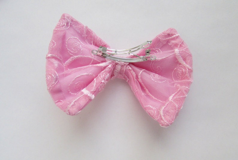 May include: A pink lace bow with a silver metal clip. The bow is made of delicate fabric with a floral pattern.