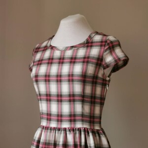 Puede incluir: Un vestido blanco con estampado de cuadros rosa y negro. El vestido tiene un escote redondo y mangas cortas. El vestido est&aacute; ajustado a la cintura y tiene una falda completa.