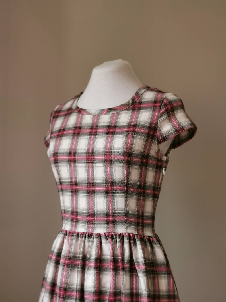 Puede incluir: Un vestido blanco con estampado de cuadros rosa y negro. El vestido tiene un escote redondo y mangas cortas. El vestido est&aacute; ajustado a la cintura y tiene una falda completa.