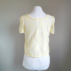 May include: A yellow lace top with short sleeves and a round neckline. The top is on a white mannequin.
