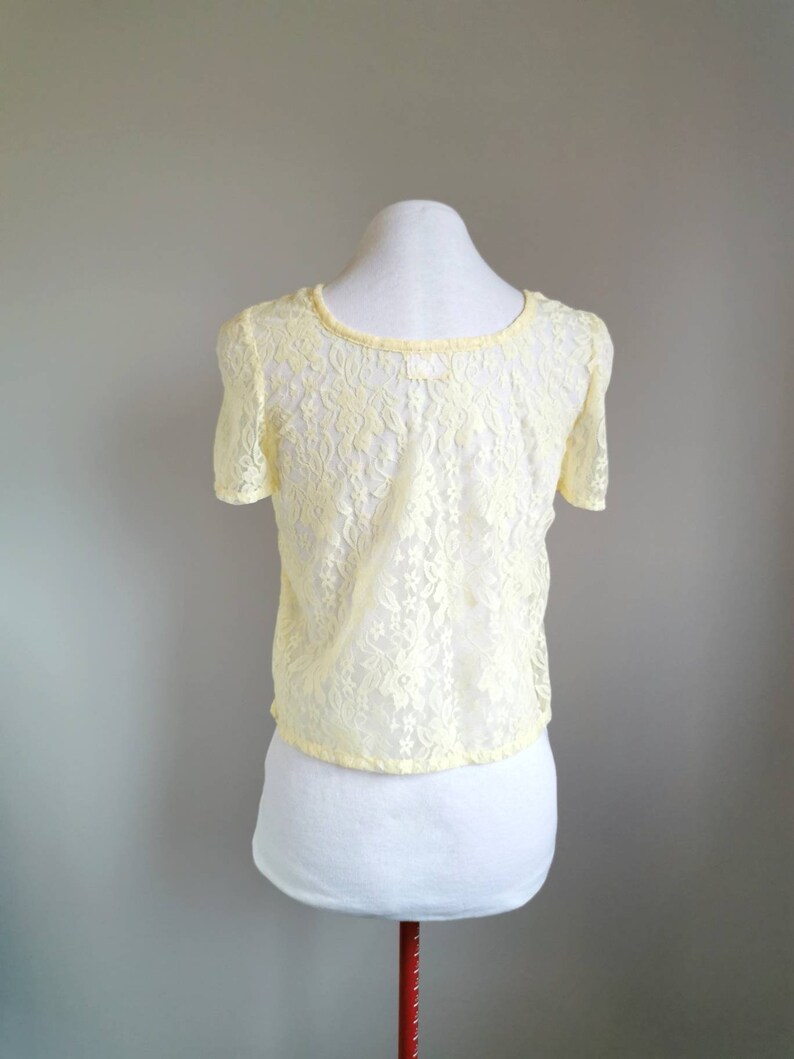 May include: A yellow lace top with short sleeves and a round neckline. The top is on a white mannequin.