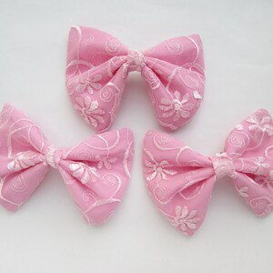 May include: Three pink fabric bows with white embroidered floral designs. The bows are tied in a classic bow shape.