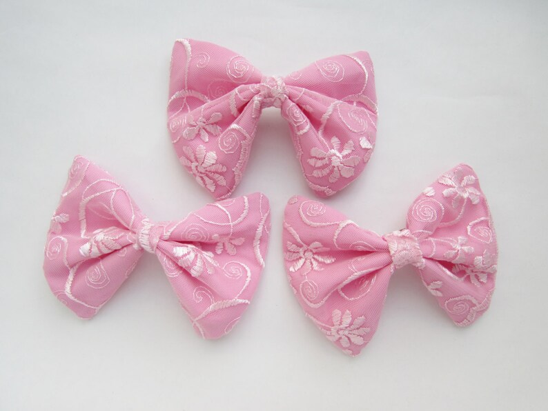 May include: Three pink fabric bows with white embroidered floral designs. The bows are tied in a classic bow shape.
