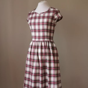 Puede incluir: Un vestido blanco con estampado de cuadros rosa y negro. El vestido tiene mangas cortas y un corpi&ntilde;o ajustado. La falda es amplia y fruncida en la cintura.