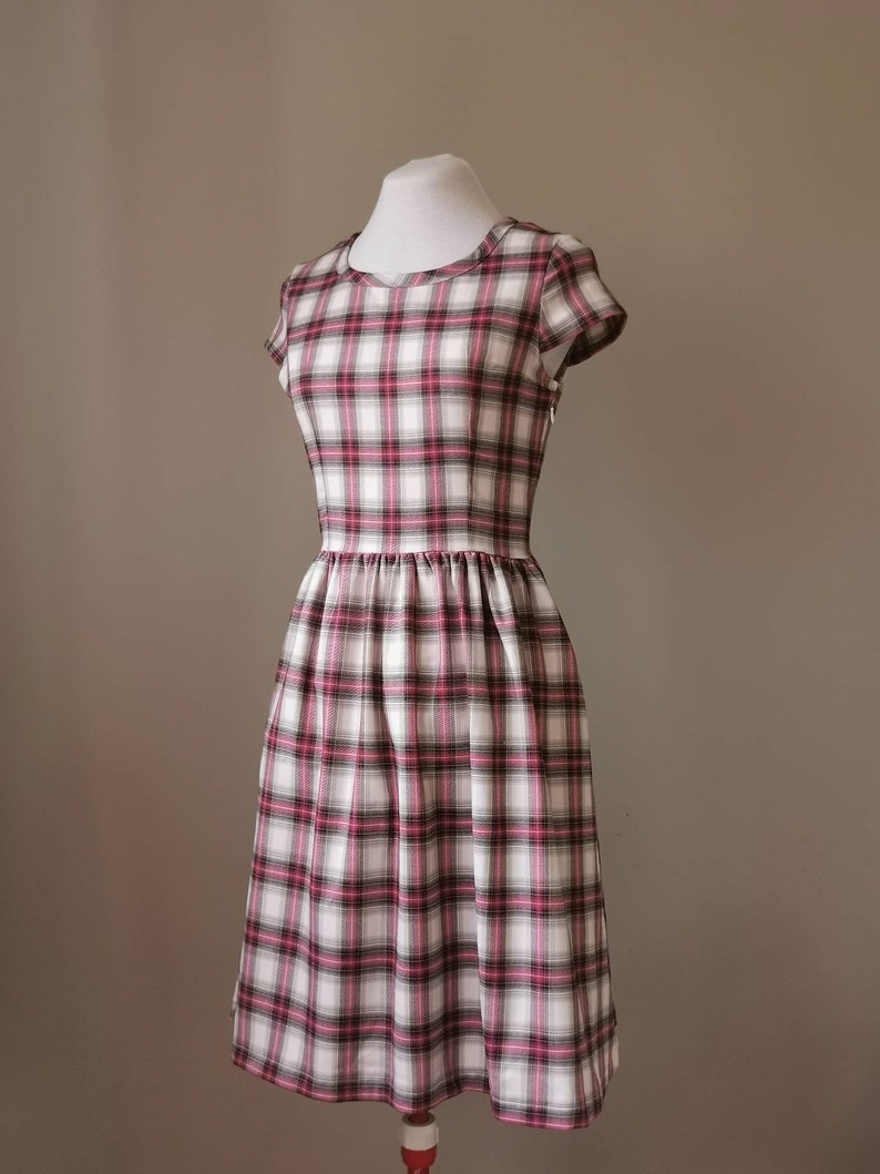 Puede incluir: Un vestido blanco con estampado de cuadros rosa y negro. El vestido tiene mangas cortas y un corpi&ntilde;o ajustado. La falda es amplia y fruncida en la cintura.