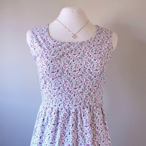 Peut inclure: Une robe blanche sans manches avec un petit imprim&eacute; floral dans les tons de violet et de vert. La robe a un col rond et un corsage ajust&eacute;. La robe est sur un mannequin blanc avec un collier en or.