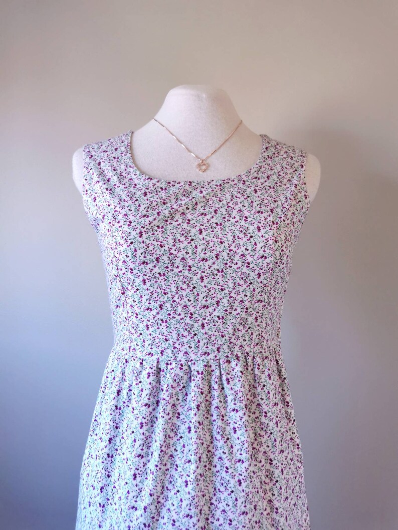 Peut inclure: Une robe blanche sans manches avec un petit imprim&eacute; floral dans les tons de violet et de vert. La robe a un col rond et un corsage ajust&eacute;. La robe est sur un mannequin blanc avec un collier en or.