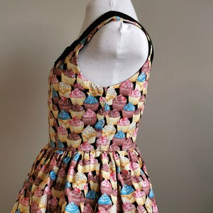K&ouml;nnte beinhalten: Ein schwarzes und gelbes &auml;rmelloses Oberteil mit Cupcake-Druck. Das Oberteil hat einen figurbetonten Oberk&ouml;rper und einen gerafften Rock.
