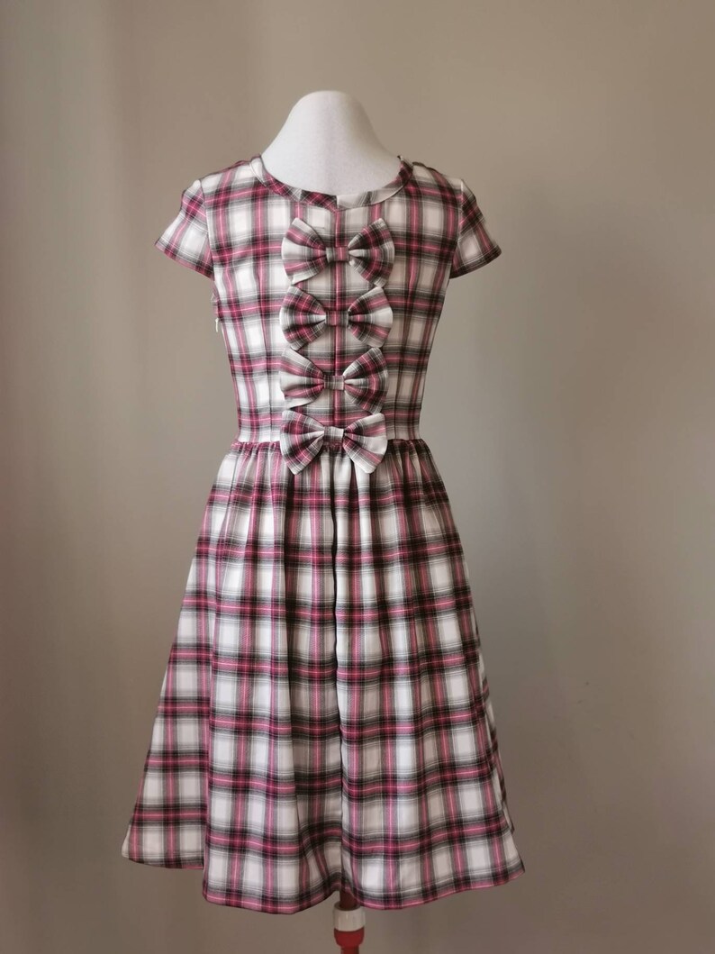 Puede incluir: Un vestido de cuadros blancos, rosas y negros con manga corta y un detalle de lazo en la parte delantera.