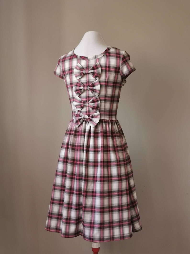 Puede incluir: Un vestido de cuadros blancos y rosas con un corpi&ntilde;o ajustado y una falda completa. El vestido tiene tres grandes lazos en la espalda, hechos del mismo tejido.