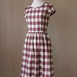 Puede incluir: Un vestido blanco con estampado de cuadros rosa y negro. El vestido tiene un escote redondo, mangas cortas y una falda fruncida.