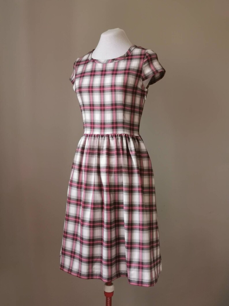 Puede incluir: Un vestido blanco con estampado de cuadros rosa y negro. El vestido tiene un escote redondo, mangas cortas y una falda fruncida.