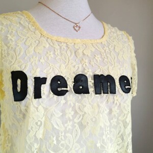 May include: A yellow lace top with short sleeves and a black embroidered "Dreamer" graphic. The top is on a white mannequin with a gold chain necklace with a small heart pendant.