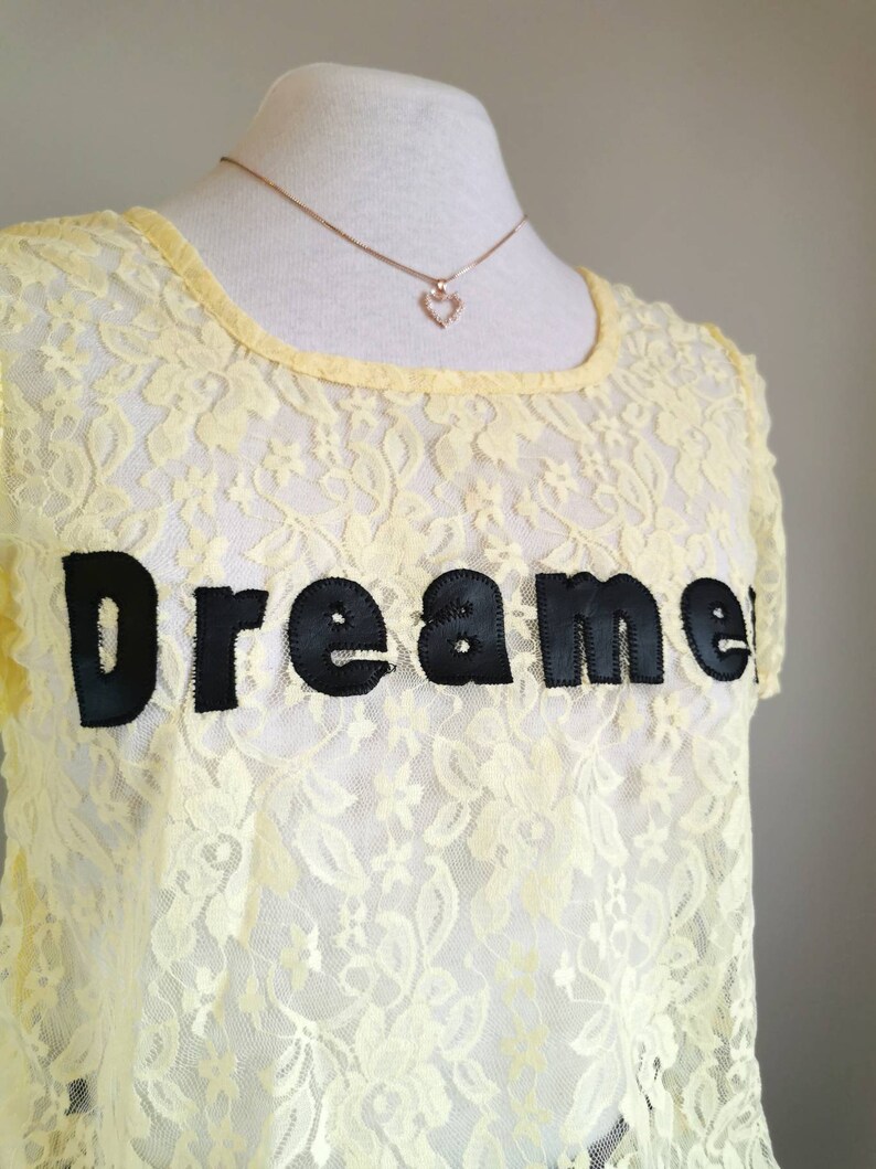 May include: A yellow lace top with short sleeves and a black embroidered "Dreamer" graphic. The top is on a white mannequin with a gold chain necklace with a small heart pendant.