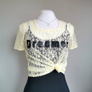 May include: A yellow lace top with a black "Dreamer" graphic. The top is tied at the front and has a scoop neckline.