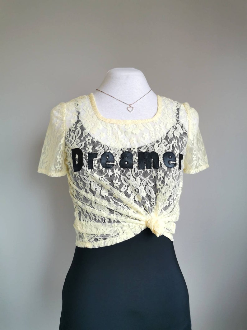 May include: A yellow lace top with a black "Dreamer" graphic. The top is tied at the front and has a scoop neckline.