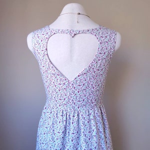 Peut inclure: Une robe blanche sans manches avec une d&eacute;coupe en forme de c&oelig;ur dans le dos. La robe a un motif floral dans les tons de violet, bleu et vert.